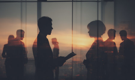 This image features silhouetted figures engaged in conversation near a window. The scene is bathed in warm hues of a sunset, suggesting an outdoor environment. The composition employs natural lighting and shadow play. Suitable for use in a variety of commercial or editorial applications related to business concepts.の素材
