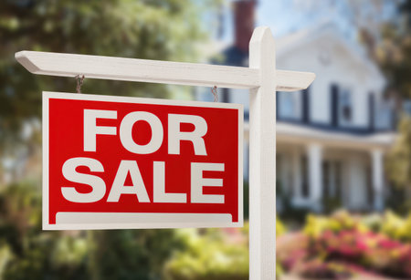 A red and white "For Sale" sign is prominently displayed in front of a house. The image showcases a bright sign with bold lettering. The background shows a blurred house with trees. Ideal for promotional material related to real estate, property listings, or related commercial use.の素材