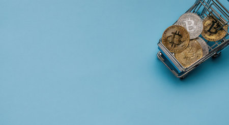 A miniature shopping cart filled with Bitcoin coins is positioned against a solid blue backdrop. The coins, exhibiting metallic tones, are in sharp focus within the cart. The composition employs a clear overhead perspective, and the lighting is even. It suits various financial themes and could be useful in illustrating economic concepts.の素材