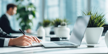 A close-up captures a person writing in a notebook near a laptop. The scene includes a blurred figure and greenery, suggesting an office environment. Soft lighting and neutral tones create a professional atmosphere. Suitable for various business, communication, and workplace-related projects.の素材