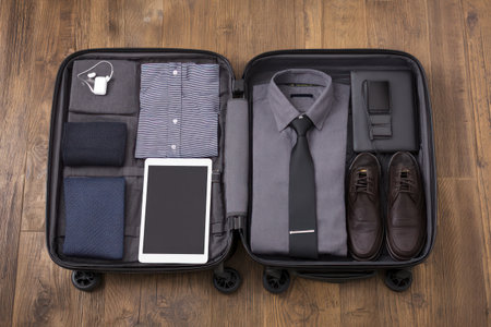 An overhead view reveals an open suitcase filled with neatly arranged clothing and accessories. It features a folded shirt, a tie, shoes, a tablet, and other personal items. The composition has a clean, organized look with a focus on details. It could be useful for travel related projects or commercial purposes.の素材