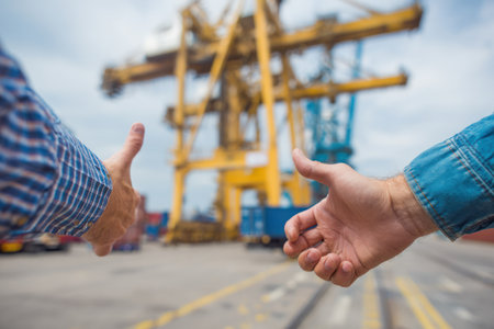 Two individuals are seen giving thumbs up, their hands prominently displayed against a blurred background. The image presents a daytime scene with a large industrial crane and shipping containers visible. The color palette includes blues, yellows, and grays, indicative of an outdoor, industrial environment. This image could be used for illustrating concepts like agreement and logistics.の素材