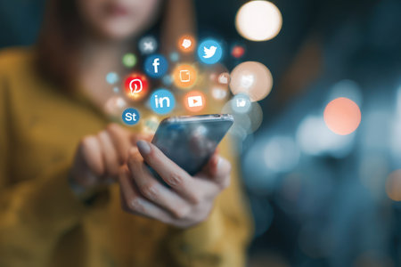 A person holds a smartphone, with colorful social media icons floating above. The image displays a shallow depth of field, with soft focus on the background. Warm tones and bokeh effects contribute to the visual appeal. Suitable for articles or websites concerning communication.の素材