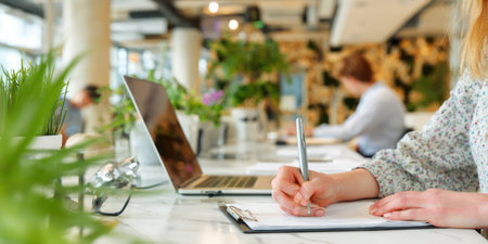 A woman is writing on a paper at a desk, near a laptop and plants. The image displays a bright, daytime setting with a shallow depth of field, blurring the background. This visual could be used in various commercial contexts, such as articles or advertising. Soft natural lighting adds warmth to the scene.の素材