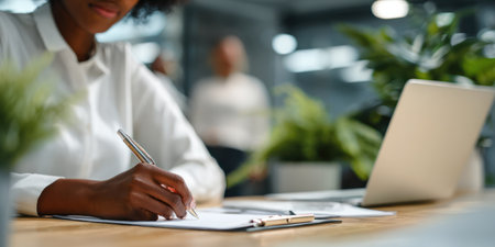 An individual is shown writing with a pen on paper, in a workspace setting. The composition includes a laptop and green plants, indicating an office or similar indoor environment. The image uses natural lighting, and offers potential use for business, education, or lifestyle related visual content.の素材