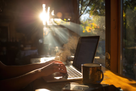 An individual uses a laptop indoors near a window with strong sunlight. The scene includes a coffee mug. The composition is a medium shot with a blurred background suggesting a room with natural lighting. This image may be suitable for illustrating concepts of remote work or creative environments.の素材