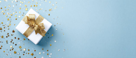 An overhead shot reveals a white gift box adorned with a golden ribbon, set against a soft blue background scattered with star-shaped confetti. The composition showcases a clean and modern aesthetic, potentially suitable for various commercial or editorial applications. The image suggests celebration and joy, potentially usable for announcements.の素材