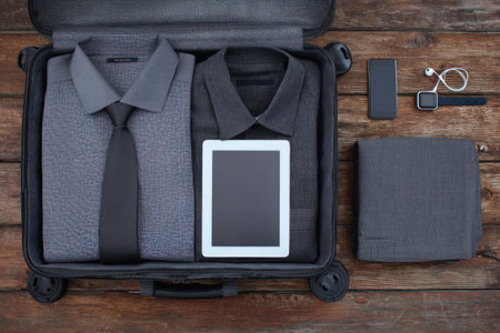 An open suitcase displays neatly folded shirts, a tie, and a tablet, accompanied by a wallet, headphones, and a smartphone. The composition features a symmetrical arrangement of gray items. Overhead lighting casts soft shadows on a weathered wooden surface. This image is suitable for illustrating travel, business, or technology themes.の素材