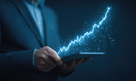 A person in a suit holds a tablet with a glowing graph depicting upward financial progress. The image features shades of blue, with bright lines creating a visual representation of growth. Suitable for concepts related to business, finance, and technology, this image may be used for various commercial projects.の素材