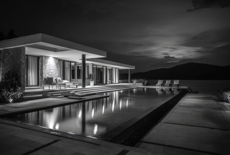 A grayscale photograph captures a modern home featuring a swimming pool, reflecting the dark sky. The composition emphasizes clean lines, geometric shapes, and a minimalist style. The lighting suggests an evening or nighttime scene with illuminated architectural details. This image is suitable for architectural, lifestyle, or travel-related content.の素材