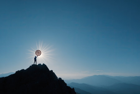 A silhouette of a person stands atop a mountain, holding a target against a bright sun. The image features a dark, rugged foreground contrasted with a clear, blue sky. The composition emphasizes achievement, aiming, and potential. Suitable for use in various business and motivational contexts.の素材