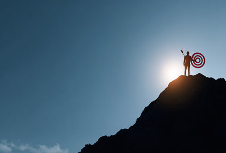 A silhouette of a person standing atop a mountain, holding a target. The image showcases a dark silhouette against a backdrop of a bright sun and a gradient blue sky. The composition emphasizes ambition and achievement. Suitable for business, motivational, and illustrative purposes.の素材