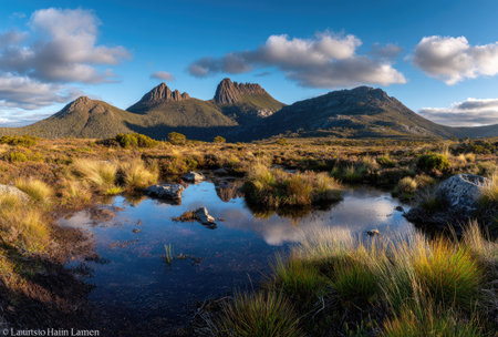 A scenic landscape showcases a mountain range mirrored in still water. The composition highlights natural textures with grassy foreground and rugged peaks. The scene is bathed in daylight under a partly cloudy sky. Suitable for environmental themes and scenic content, offering visual appeal.の素材