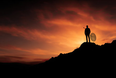 A silhouette depicts a person standing on a hill, holding a target against a dramatic sunset. The image displays deep orange and red hues in the sky, creating a contrasting background. The composition suggests determination and goal setting, suitable for various promotional and illustrative projects.の素材