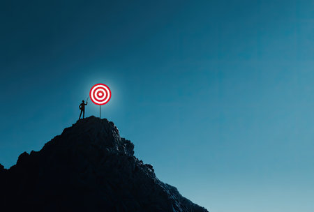 A silhouette of a person stands atop a mountain, aiming towards a glowing target. The image showcases a dark, textured rock formation against a bright, gradient blue sky. The composition is simple, using an overhead shot. It may be suitable for illustrating concepts of achievement and success.の素材