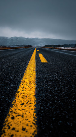 An asphalt road, marked with prominent yellow lines, stretches into the distance, framed by a cloudy sky and mountains. The image displays a strong vertical composition, featuring dark tones, emphasizing the path. This image is suitable for various projects requiring visuals related to travel or journey, and design layouts.の素材