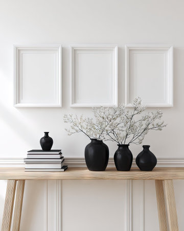 A minimalist interior arrangement features three empty picture frames above a wooden table. The table displays decorative black vases and a stack of books. The composition employs a neutral color palette with white walls, natural wood, and black accents, suitable for various design projects and commercial applications.の素材