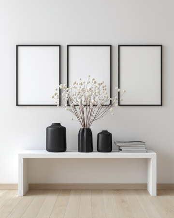 A minimalist interior setting showcases three blank picture frames above a white console. The composition features a delicate floral arrangement in black vases, complemented by stacked books. The scene displays a neutral color palette, employing natural light, ideal for commercial projects needing a modern or stylish presentation.の素材