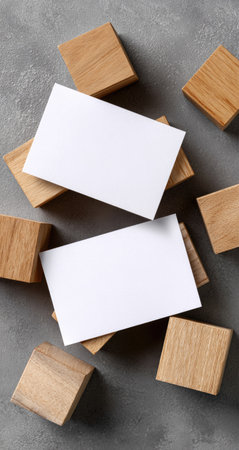 Two white business cards rest on top of several wooden blocks arranged on a textured gray surface. The overhead shot showcases a minimalist composition with natural light and shadow play. Suitable for presentations, advertising, and editorial projects, it offers copy space for text or design elements.の素材