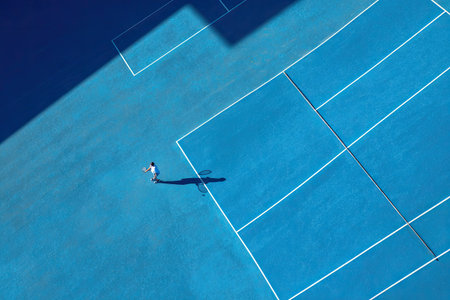 An overhead shot reveals a bright blue tennis court with white lines and a player in action. The scene is illuminated by sunlight, creating a stark contrast between light and shadow. The composition highlights the geometric pattern of the court. Suitable for sports publications, marketing material, and various commercial purposes.の素材