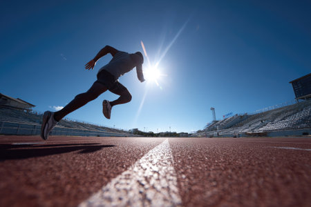 An athlete is captured mid-stride, sprinting on a red track with a prominent white line. The composition features a low-angle shot, highlighting the runner's silhouette against a clear blue sky and the bright sun. The image conveys motion, energy, and determination, potentially suitable for promotional or editorial purposes.の素材