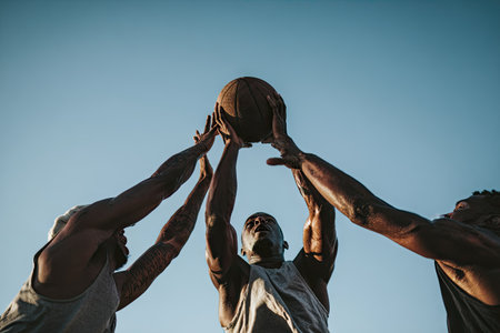 Three individuals are reaching for a basketball against a backdrop of a bright, cloudless sky. The players are seen from a low angle, emphasizing the upward reach. The composition features a neutral color palette. This image could be used for various commercial or editorial applications.の素材