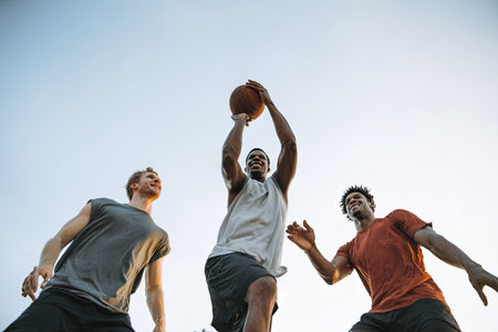 Three individuals are captured mid-action, focused on a basketball held high. They are wearing athletic apparel under a clear, pale-blue sky. The image has a low-angle perspective, emphasizing motion and effort. This photograph could be used for sports articles, training publications, or promotional material.の素材