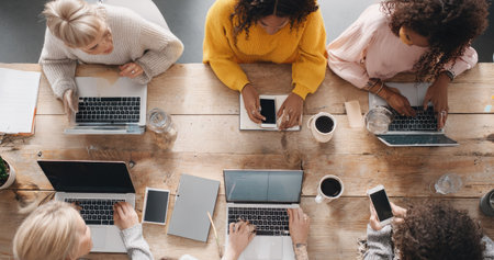 An overhead view reveals a diverse group of women working together at a wooden table. They are using laptops and smartphones, possibly engaged in collaborative tasks. The scene is bright, with soft lighting and a focus on technology. This image could be used for various commercial or editorial projects related to teamwork and technology.の素材