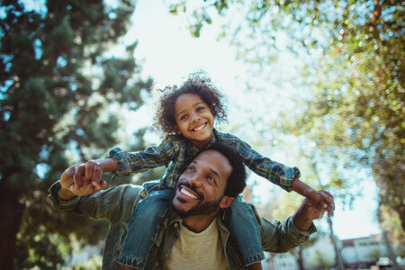 A man carries a young child on his shoulders, both smiling. The image showcases natural light and a soft focus. The composition emphasizes the relationship between the two individuals. This photo could be suitable for family-related content and other commercial applications.の素材
