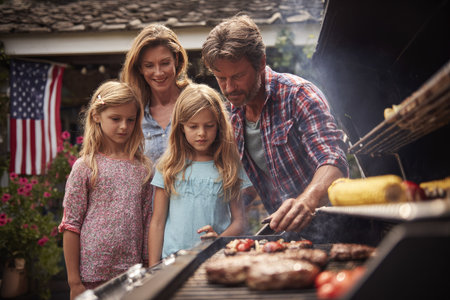 A family gathers around a barbecue grill, preparing a meal outdoors. The scene features a man and woman with their two daughters, with an American flag visible in the background. The lighting suggests a sunny day, with vibrant colors and textures adding visual appeal. This image is suitable for various commercial uses, including lifestyle and family-oriented content.の素材