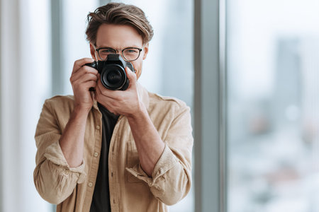 A man wearing glasses holds a camera, positioned near a window. The image displays natural lighting, and a shallow depth of field. The subject is likely photographing something. This photograph could be utilized for various commercial purposes, including marketing and advertising campaigns.の素材