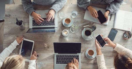 An overhead shot reveals colleagues engaged in teamwork around a table, utilizing laptops, tablets, and smartphones. The scene includes cups of coffee and various objects on a wooden surface. The composition has an even lighting and neutral tones, suitable for business or informational uses.の素材
