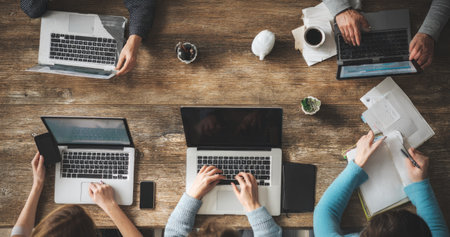 This overhead shot captures a collaborative work session. Several people are gathered around a wooden table, using laptops and taking notes. The composition shows hands, devices, and office supplies. The warm lighting and natural texture suggest an indoor setting, suitable for business or editorial use.の素材