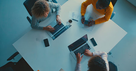 An overhead perspective showcases four individuals gathered around a white table, each focused on a laptop. The composition highlights teamwork and technology use. The scene is brightly lit, with a modern aesthetic, suggesting an office or collaborative setting. Suitable for various applications related to business or technology.の素材