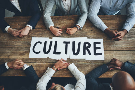 Several people in professional attire gather around a wooden table with a sign reading "CULTURE." The overhead shot showcases a diverse group. The scene suggests a meeting or discussion within a collaborative environment with natural lighting. This image could be used for business presentations, articles, or various editorial contexts.の素材
