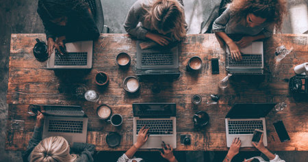 An overhead view reveals individuals engaged in collaborative work around a rustic wooden table. The composition highlights laptops, cups, and personal items. The scene is illuminated by natural light, creating a warm atmosphere suitable for use in business and lifestyle publications.の素材