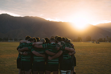 A rugby team gathers closely together in a huddle on a green field at dusk. The players, wearing uniforms, have their arms around each other. The scene is bathed in warm light, with the sun setting behind distant mountains. This image could be used for sports-related articles, team building, or motivational content.の素材