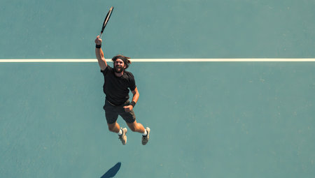 An aerial perspective shows a tennis player mid-air on a court. The player, dressed in sportswear, is reaching upwards with a racket. The scene features a teal court with white lines, suggesting an outdoor environment with ample daylight. The image may be suitable for athletic promotions, editorial content, or sports-related applications.の素材