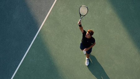 A tennis player performs an overhead serve on a green court. The image shows the player from an aerial perspective. The composition emphasizes the player's action and the court's geometry. The scene is bathed in sunlight, enhancing the colors. This image is suitable for various commercial uses related to sports and fitness.の素材