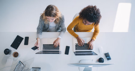 Two women are shown working at a desk using laptops, viewed from an overhead perspective. The scene exhibits natural lighting. The overall composition suggests a business or collaborative environment, useful for various commercial and editorial projects requiring a visual of teamwork and technology.の素材
