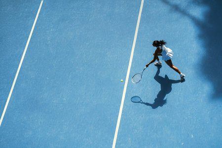 An overhead shot captures a tennis player in mid-swing on a bright blue court. The image shows a woman in athletic wear, casting a long shadow under direct sunlight. The composition emphasizes lines and negative space, suitable for commercial projects promoting sports and fitness.の素材