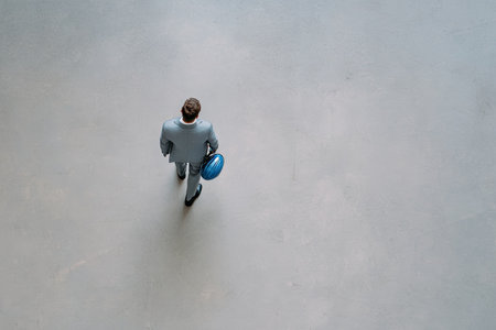 A man in a suit is seen walking across a blank, light-colored surface. The overhead shot reveals a minimalist composition, featuring the figure carrying a blue object. The image is rendered with a neutral tone, providing ample copy space, suitable for various business or conceptual applications.の素材