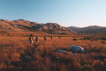A group of people are seen walking through a wide open, grassy field during the day. The scene shows a dry, warm environment with brown vegetation and a distant mountain range under a clear, light blue sky. This image may be suitable for illustrating themes of travel and exploration.の素材
