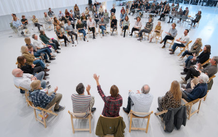 An overhead view displays a large group of people arranged in a circular formation, seated on chairs. The composition features a bright, white floor. The individuals appear to be engaged in conversation, with some raising their hands. This image is suitable for various commercial uses, including illustrating concepts of community and collaboration.の素材