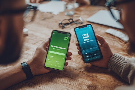 Two individuals are shown holding smartphones, displaying applications with user interfaces. The image presents a close-up perspective, focusing on the devices and hands. The environment features a wooden table, glasses, and a laptop suggesting a collaborative work setting. Suitable for various business, technology, and communication related projects.の素材