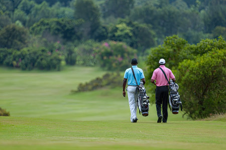 Two individuals are depicted walking on a well-maintained grassy golf course, each carrying a golf bag. The scene showcases green foliage and trees in the background, suggesting an outdoor setting during daylight. The image may be suitable for editorial or commercial content related to sports and leisure activities.の素材