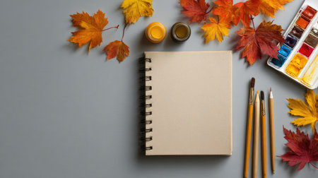 An overhead shot presents a creative workspace featuring a blank notebook alongside paint supplies and autumn leaves. The composition showcases a neutral gray surface, with vibrant maple leaves of red, orange, and yellow hues. The arrangement suggests an environment for art, design, or decorative projects. The image can be suitable for editorial or commercial purposes.の素材