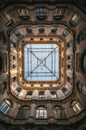 An architectural interior presents an upward perspective of an atrium with a large glass skylight. The composition highlights repeating windows and structural elements. The color palette emphasizes warm tones with highlights and shadows, giving depth. Ideal for illustrating construction, design, or background use.の素材