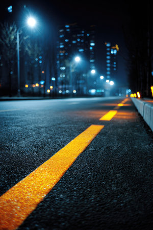 A close-up view presents an asphalt road with a prominent yellow line, illuminated by streetlights and building lights. The composition highlights contrasting textures and colors, from the dark road to the bright lights. This image could be used for various commercial or editorial purposes related to urban themes or nighttime scenes.の素材