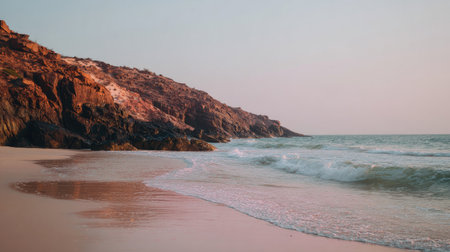 The image depicts a tranquil coastal scene with gentle waves lapping against a sandy shore. A rocky cliff is present to the left. Warm colors dominate the composition, suggesting a serene atmosphere. Ideal for a variety of commercial applications, the image is suitable for promotional materials or editorial content.の素材